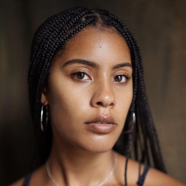 Close-up portrait of a woman with braided hair and hoop earrings looking directly at the camera