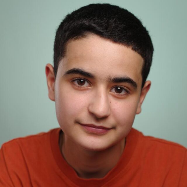 Close-up portrait of a young man with short dark hair, wearing an orange top, against a pale green background