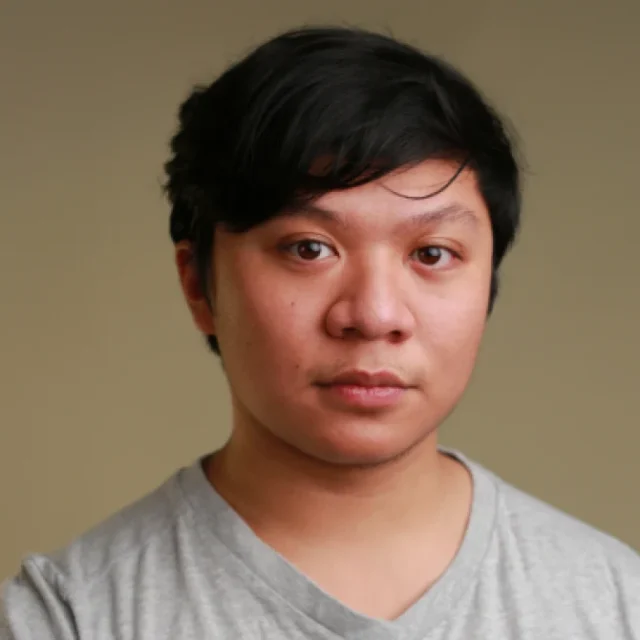 Close-up portrait of a young man with short dark hair, wearing a grey T-shirt, looking directly at the camera