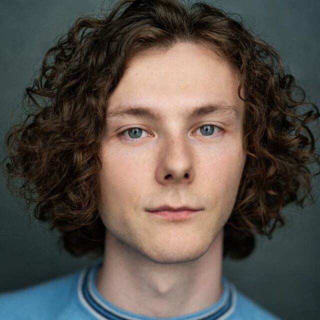 Young man with curly brown hair and blue eyes, looking directly at the camera with a neutral expression