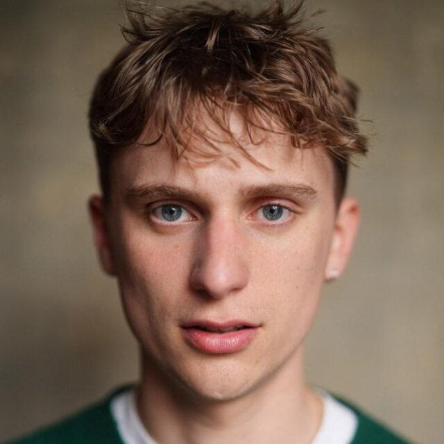 Close-up portrait of a young man with tousled brown hair and blue eyes, wearing a green top