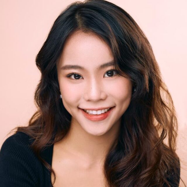 Smiling woman with long wavy dark hair, wearing a black top, against a plain pink background
