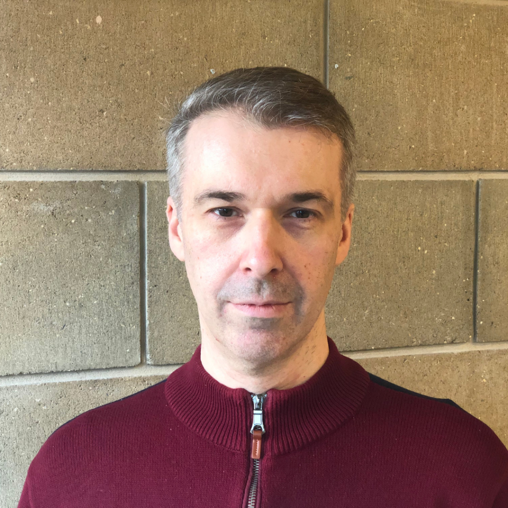 Headshot of a man with short greying hair wearing a maroon zip-up jumper, standing against a stone block wall