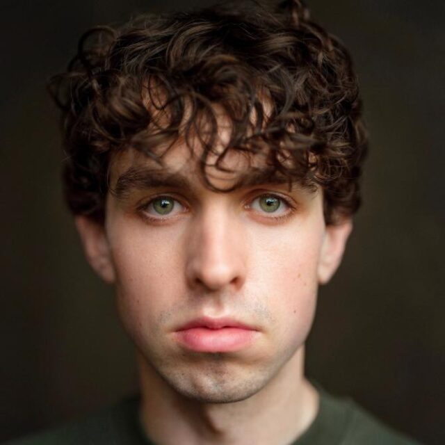 Close-up portrait of a young man with curly brown hair and green eyes, looking directly at the camera with a neutral expression