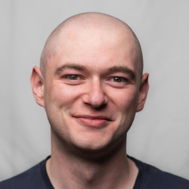 Headshot of a bald man smiling at the camera, wearing a dark T-shirt against a plain grey background