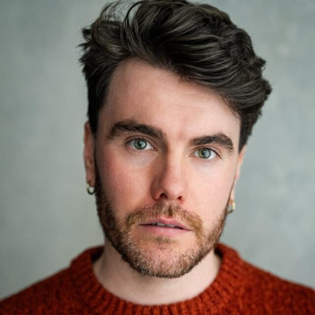 Close-up portrait of a man with dark wavy hair, a beard and earrings, wearing a rust-coloured knitted jumper