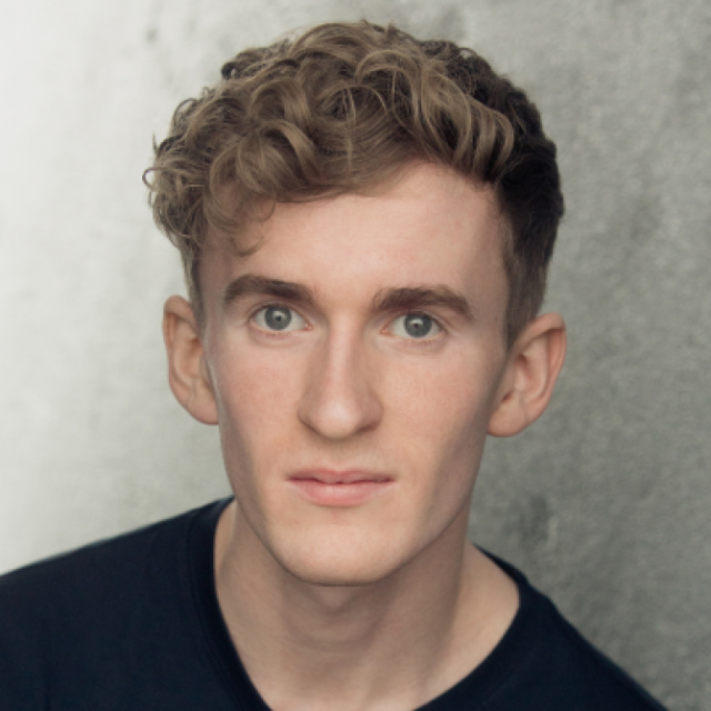 Headshot of a young man with curly light brown hair, wearing a black top, against a grey background