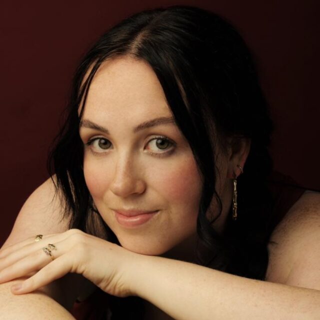 Close-up portrait of a woman with dark hair resting her chin on her hand, against a deep red background