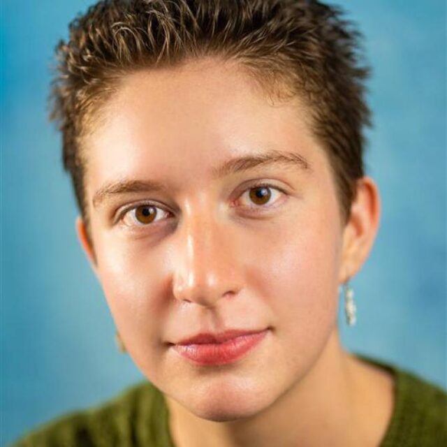 Close-up portrait of a person with short brown hair, wearing earrings and a green jumper, against a blue background