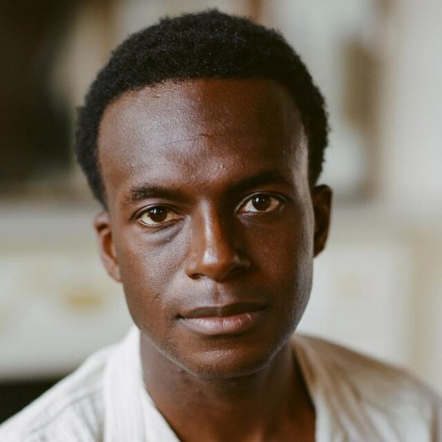 Close-up portrait of a young man with short black hair, looking directly at the camera with a neutral expression