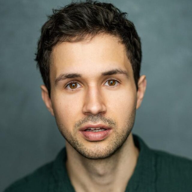 Headshot portrait of a young man with dark curly hair, wearing a dark green top