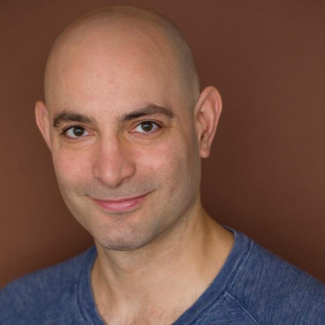 Close-up portrait of a smiling bald man wearing a blue T-shirt against a brown background
