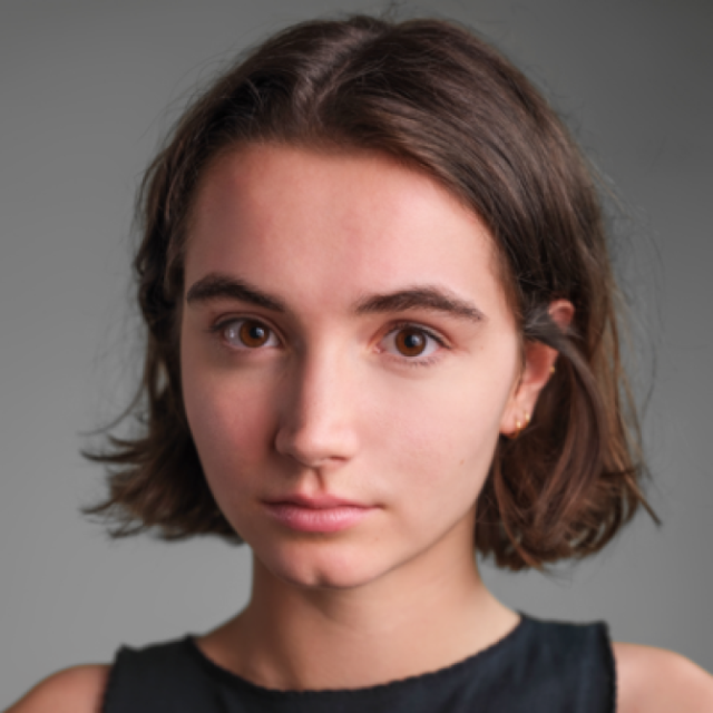 Close-up studio portrait of a young woman with shoulder-length brown hair, looking directly at the camera against a grey background