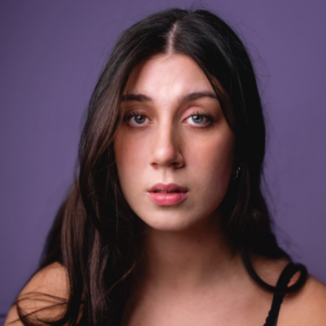 Close-up portrait of a young woman with long dark hair, looking directly at the camera against a purple background