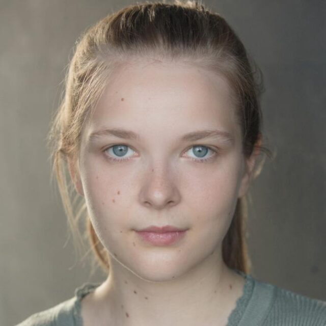 Close-up portrait of a young woman with fair skin, blue eyes and light brown hair pulled back, looking directly at the camera