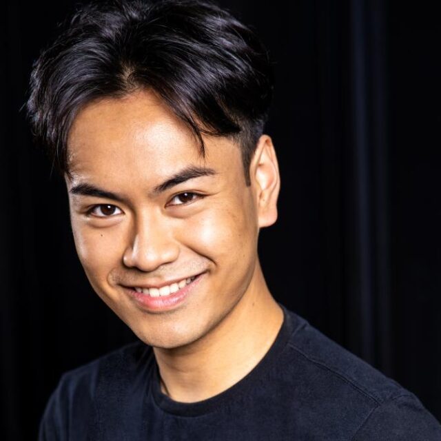 Close-up portrait of a smiling young man with dark hair, wearing a black top against a dark background