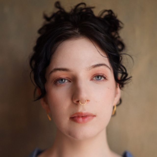 Close-up portrait of a woman with curly dark hair, blue eyes, a gold septum ring and small hoop earrings