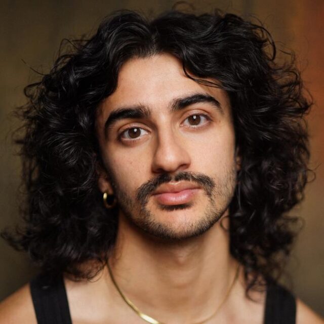 Close-up portrait of a man with long curly dark hair, a moustache and a gold necklace, looking directly at the camera