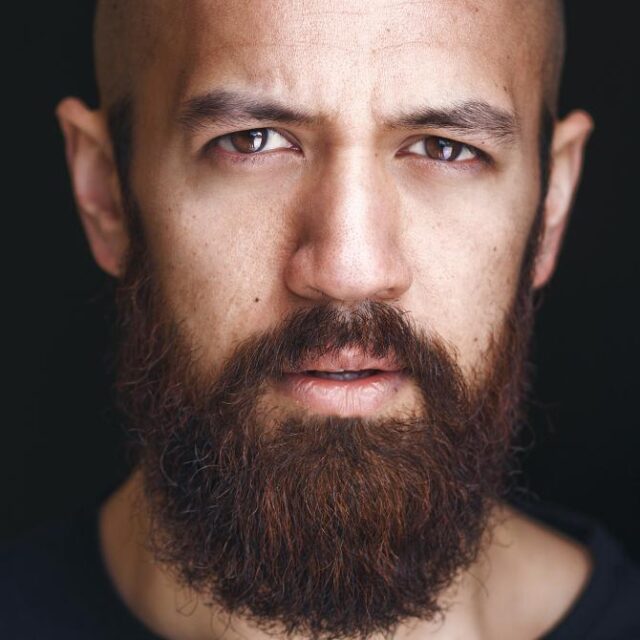 Close-up portrait of a bald man with a full reddish-brown beard, looking directly at the camera with an intense expression