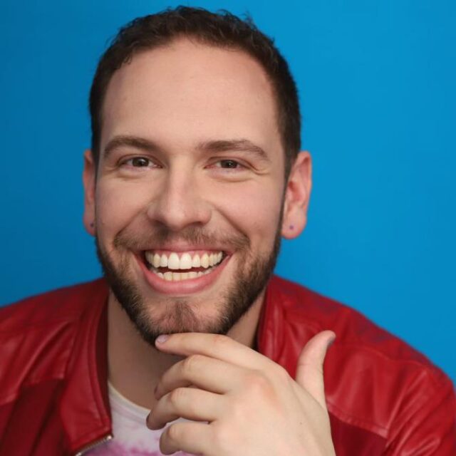 Man with a beard smiling broadly, wearing a red leather jacket against a blue background