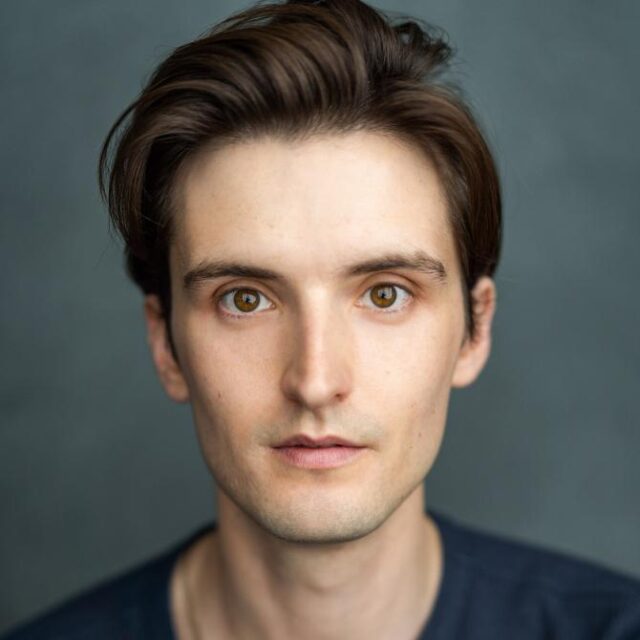 Headshot of a young man with brown hair and hazel eyes, looking directly at the camera against a grey background