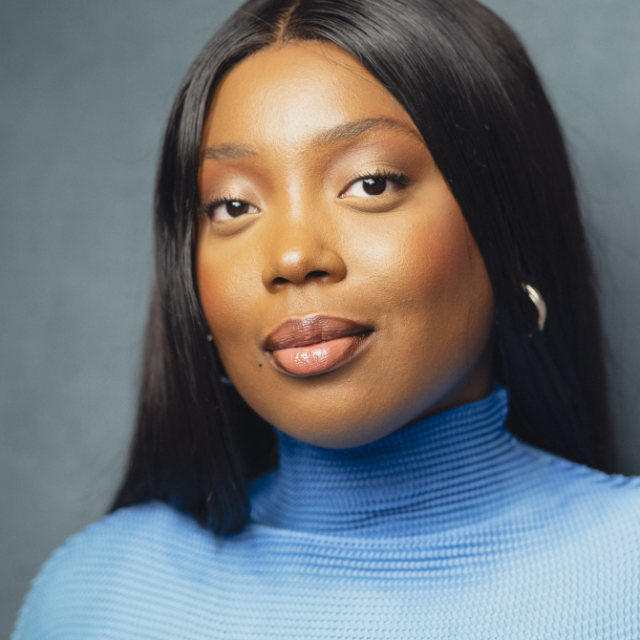 Close-up portrait of a woman with long dark hair wearing a blue polo-neck jumper, looking directly at the camera
