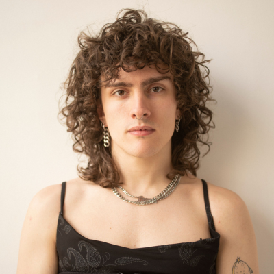 Person with curly brown hair, wearing a chunky silver chain necklace and dark patterned top, looking at the camera