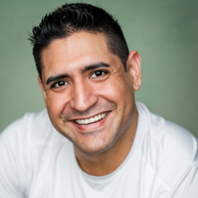 Smiling man with short dark hair and an earring, wearing a white top, against a green background