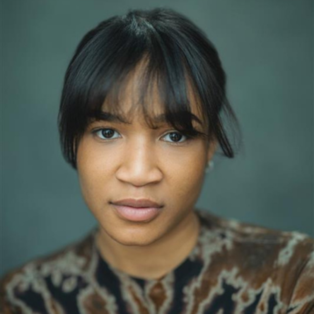 Close-up portrait of a young woman with dark hair and fringe, wearing a patterned top, against a grey background