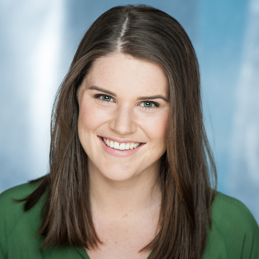Headshot of a smiling woman with long brown hair, wearing a green top, against a blue background