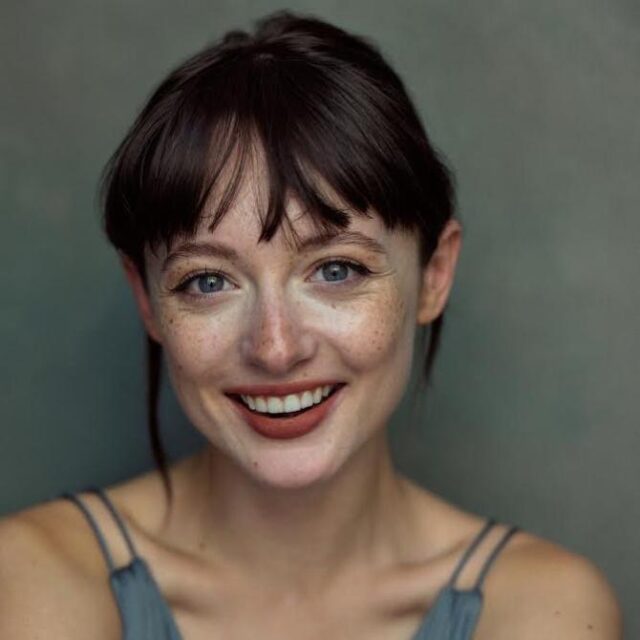 Woman with dark hair and fringe, freckles and blue eyes, smiling at the camera