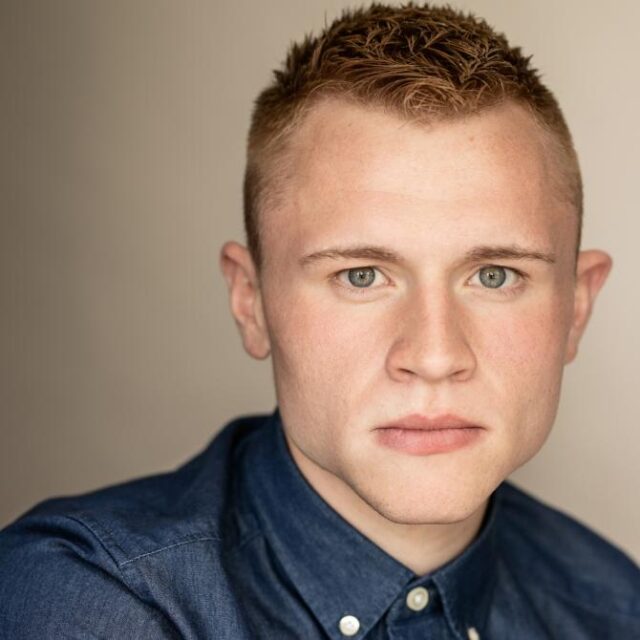 Close-up portrait of a young man with short ginger hair, wearing a denim shirt, looking directly at the camera
