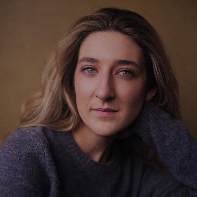 Close-up portrait of a woman with long wavy hair in a grey jumper, resting her head on her hand