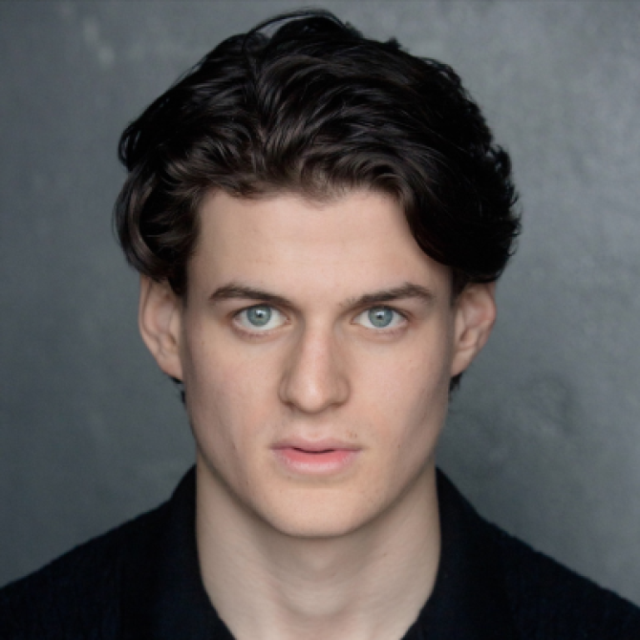 Headshot of a young man with wavy dark hair and green eyes, wearing a black top