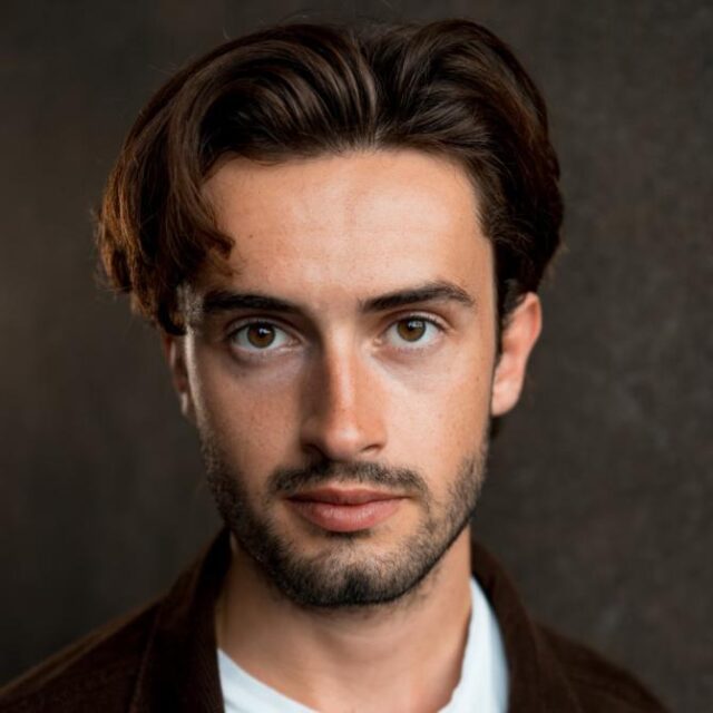 Close-up portrait of a young man with dark wavy hair and a short beard, looking directly at the camera