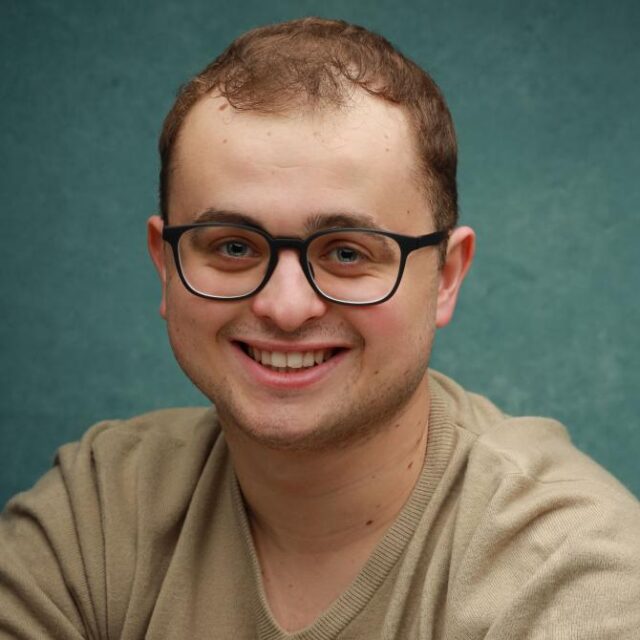 Smiling man with glasses and short brown hair, wearing a beige jumper