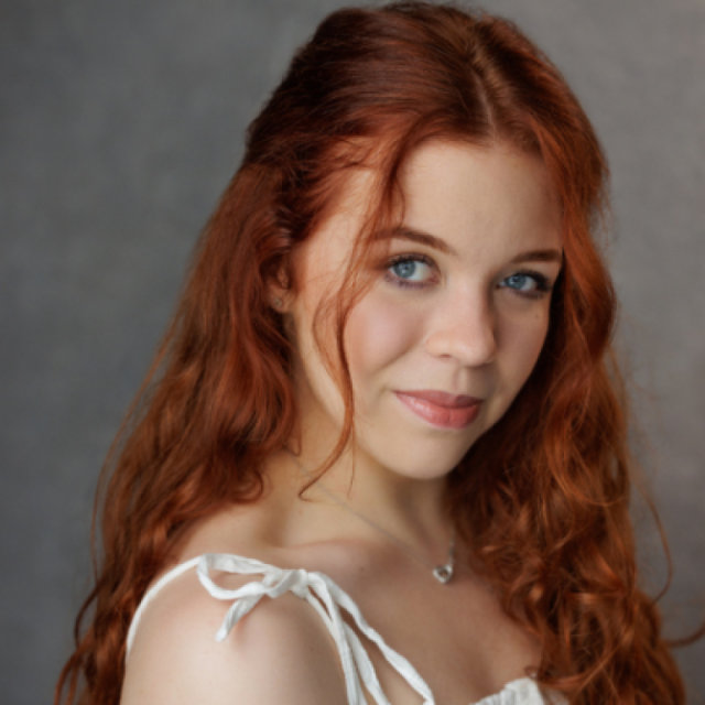Portrait of a young woman with long red curly hair, wearing a white top and a small heart pendant necklace