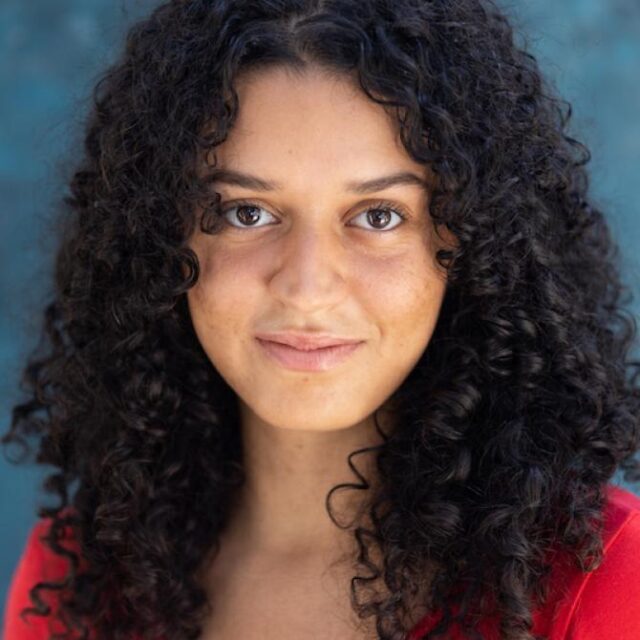 Portrait of a woman with long curly dark hair, wearing a red top, smiling gently against a blue background