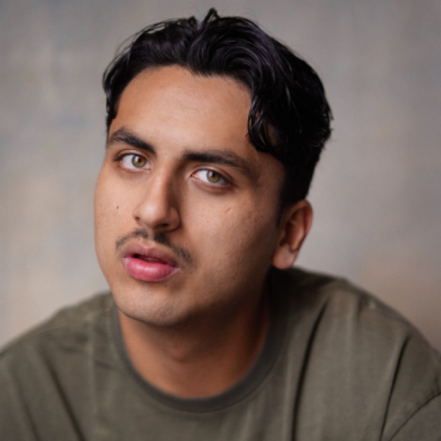 Close-up portrait of a young man with dark wavy hair and a moustache, wearing an olive green T-shirt