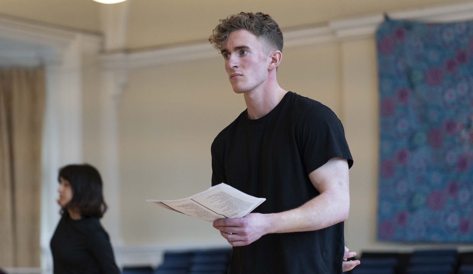 Actor holding a script during a rehearsal, looking off to the side in a bright hall