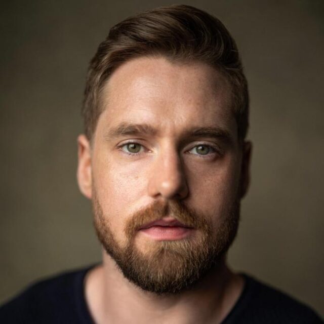 Close-up portrait of a man with short brown hair and a full beard, looking directly at the camera against a dark background