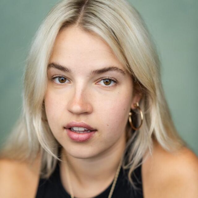 Close-up portrait of a young woman with blonde hair and gold hoop earrings, looking at the camera