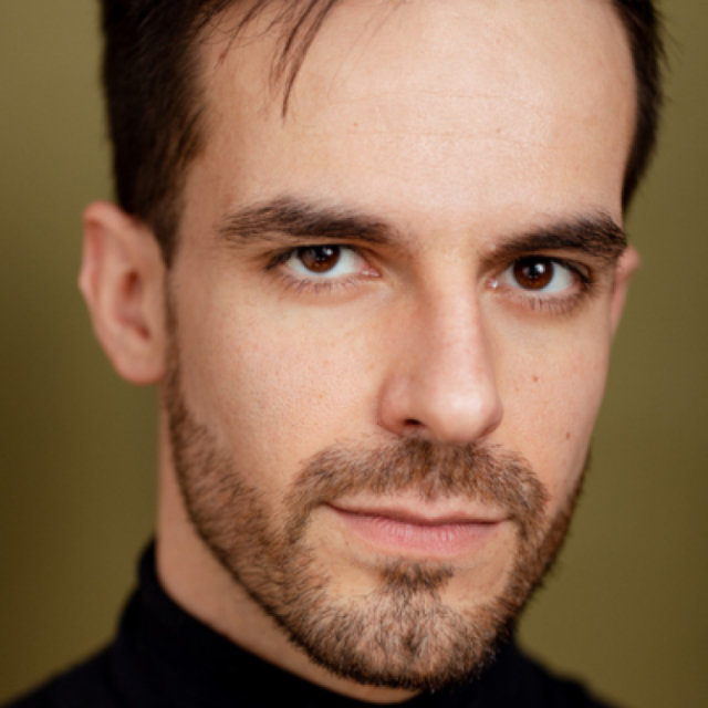 Close-up portrait of a man with dark hair and a beard, wearing a black top, looking at the camera