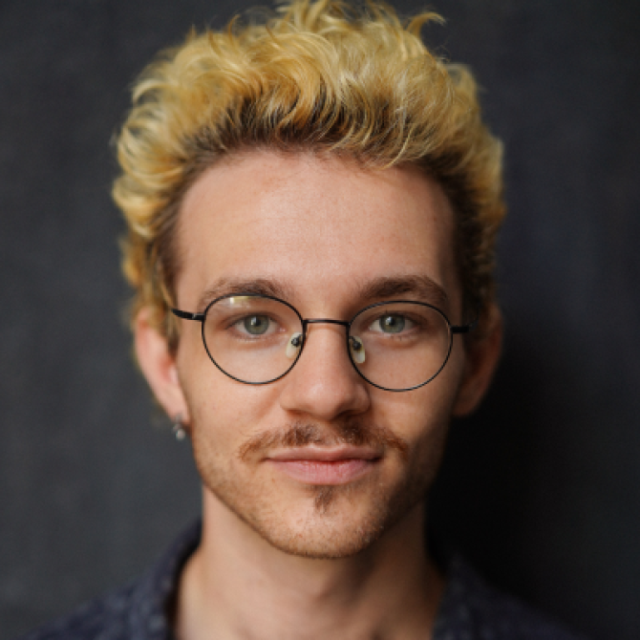 Man with curly blond hair and round glasses, smiling slightly against a dark background
