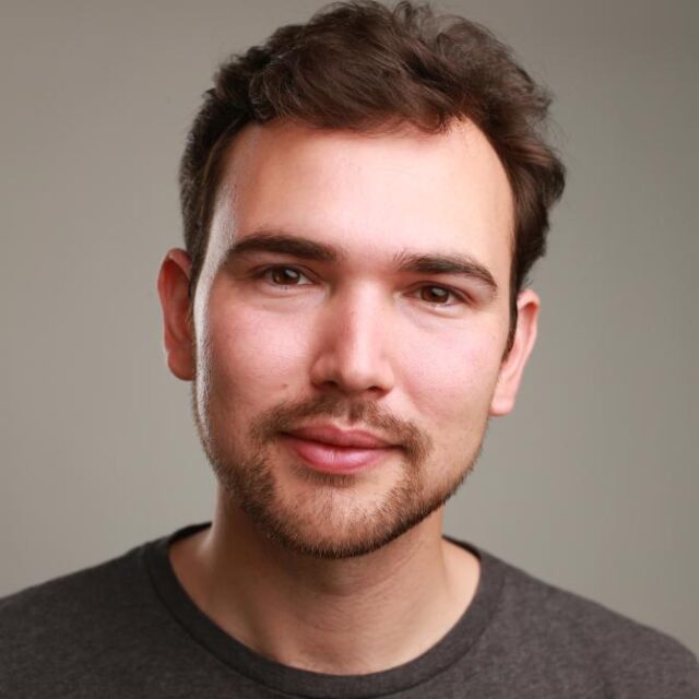 Headshot of a young man with short brown hair and stubble, wearing a dark grey top, looking directly at the camera