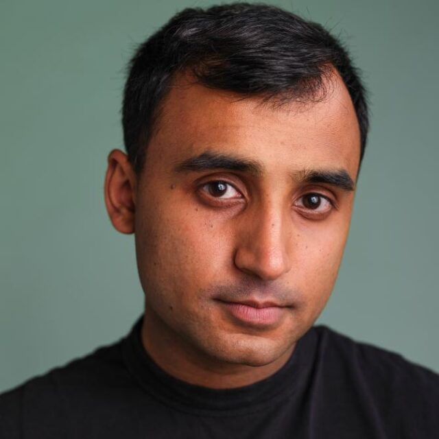 Close-up portrait of a man with short dark hair, wearing a black top, against a plain green background