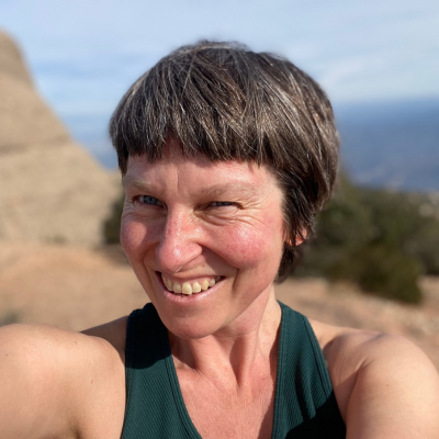 Smiling woman with short grey-brown hair taking a selfie outdoors on a rocky hillside
