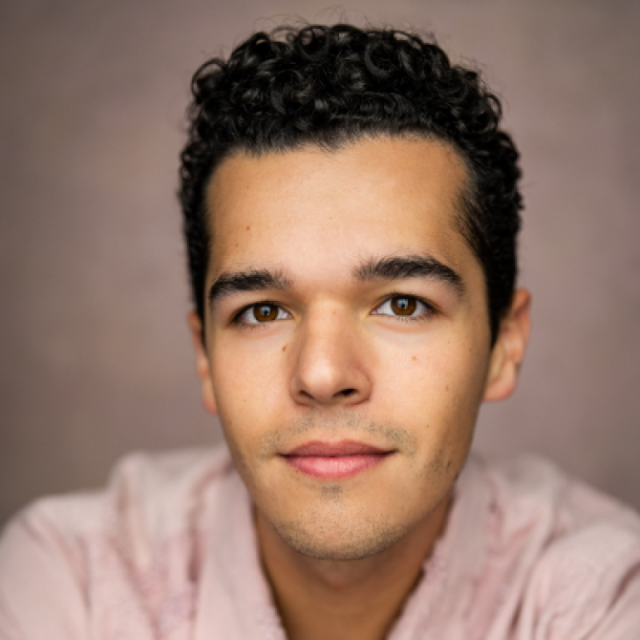 Close-up portrait of a young man with curly dark hair, wearing a pale pink shirt, looking directly at the camera