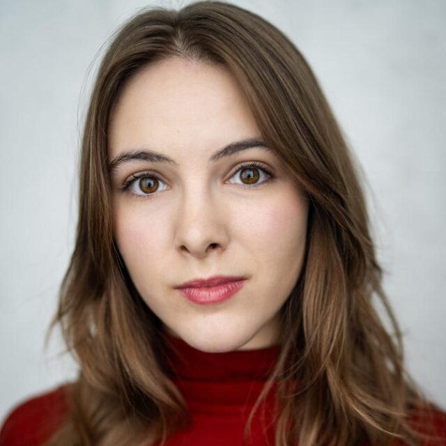 Close-up portrait of a young woman with long brown hair, wearing a red top, looking directly at the camera