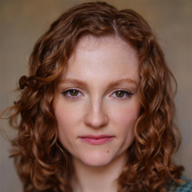 Headshot of a woman with curly auburn hair, looking directly at the camera with a neutral expression